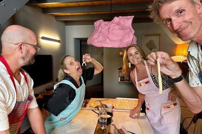 A group of people in a kitchen, some wearing aprons, with a pink cloth hanging above them. 1