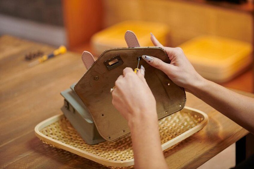 Manos armando un objeto pequeño, marrón, con apariencia de cuero y cierres metálicos sobre una mesa de madera. 2