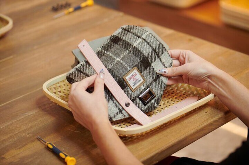 Hands assembling a woven bag with a plaid pattern and metal hardware on a wooden table. 1