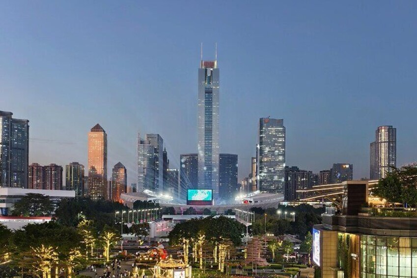 A city skyline with a prominent skyscraper, illuminated buildings, and a large digital display. 3
