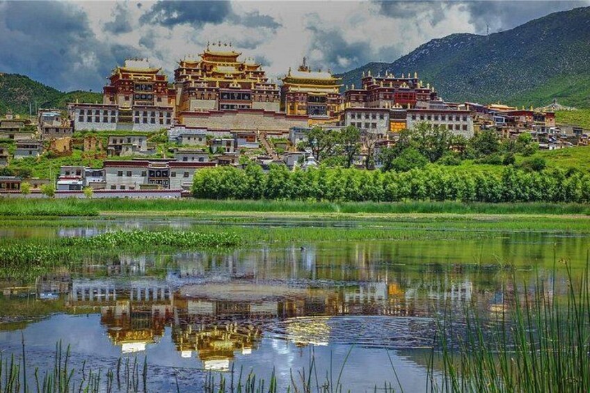 A traditional Tibetan monastery complex with multiple buildings, a reflection in a body of water, and a mountainous backdrop. 2