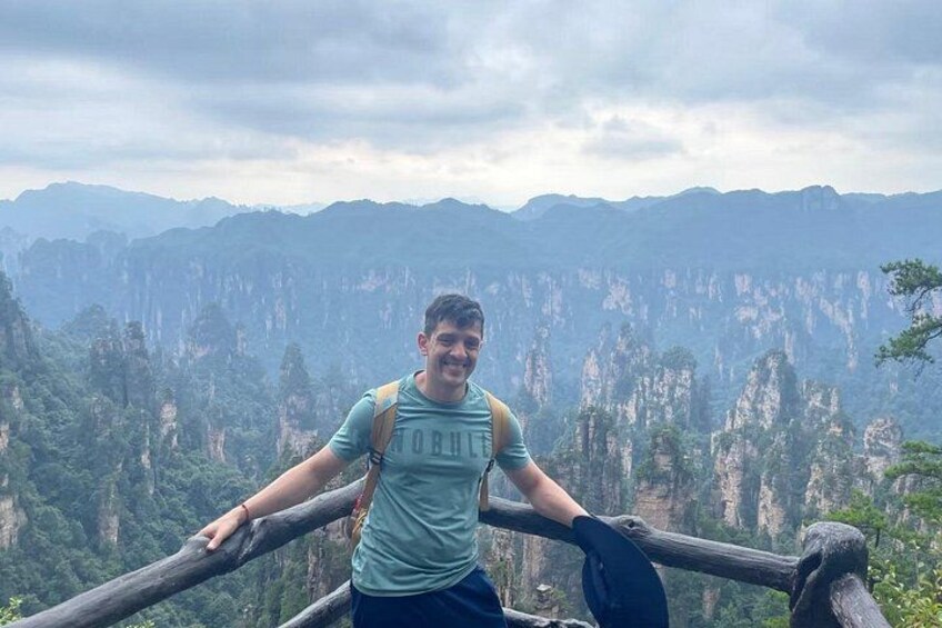 A person standing on a wooden platform overlooking a mountainous landscape with steep cliffs and dense forest. 2