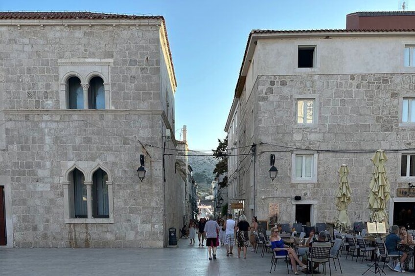 A stone building with arched windows and a tiled roof. People are walking and sitting outside. 5