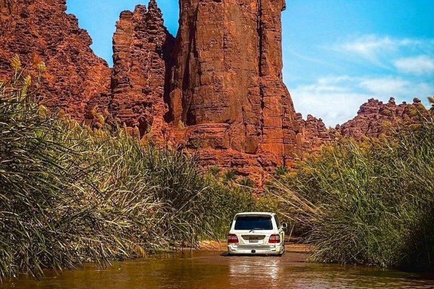 A white SUV driving through a narrow water channel surrounded by red rock formations and green vegetation. 3