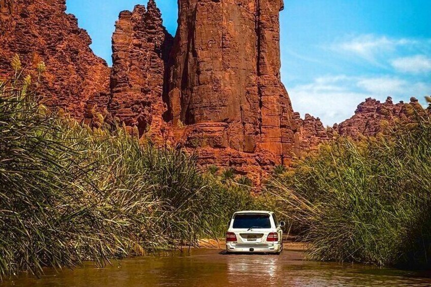 A white SUV driving through a narrow water channel surrounded by red rock formations and green vegetation. 3