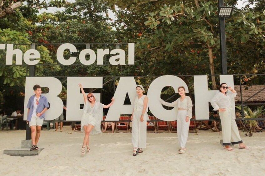 A beach with large letters spelling 'The Coral Beach' and people posing in front of it. 2