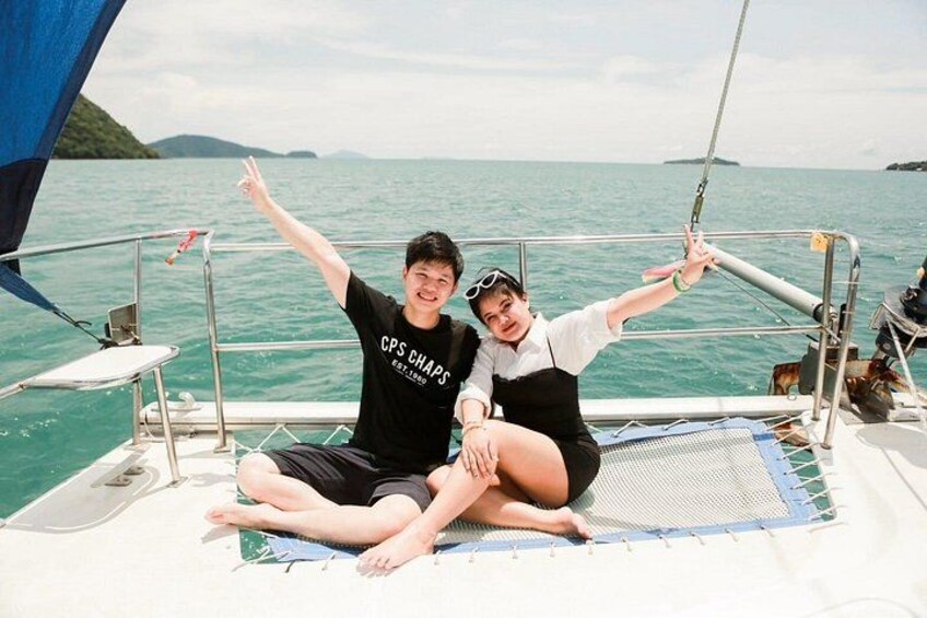 Two people sitting on a boat with a blue sail, surrounded by water and distant land. 3