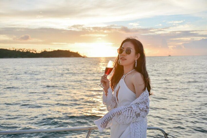 Une personne tenant un verre contenant un liquide rouge, debout sur un bateau, avec une étendue d'eau et un coucher de soleil en arrière-plan. 2