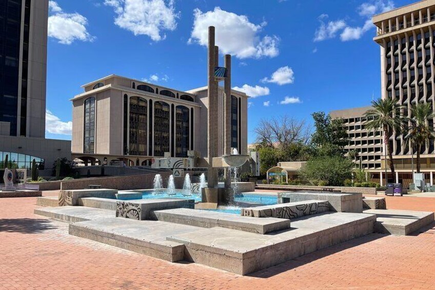 A public square with a fountain, a tall building with columns, and a smaller building with a large window. 3