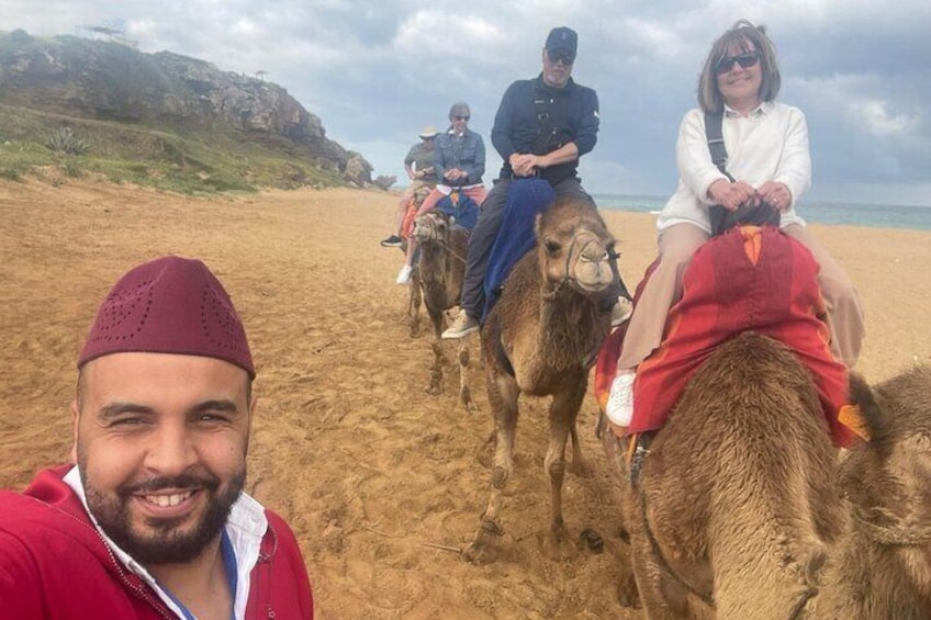 Personas montando camellos en una playa arenosa, con un paisaje rocoso al fondo. 1