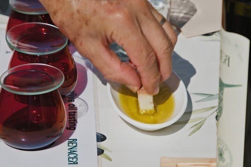 A hand dipping a piece of bread into a bowl of yellow liquid, possibly oil, with red containers in the background. 2