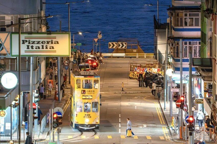 A yellow tram at a street intersection with a pedestrian crossing, a sign for 'Pizzeria Italia,' and a view of the water in the background. 2