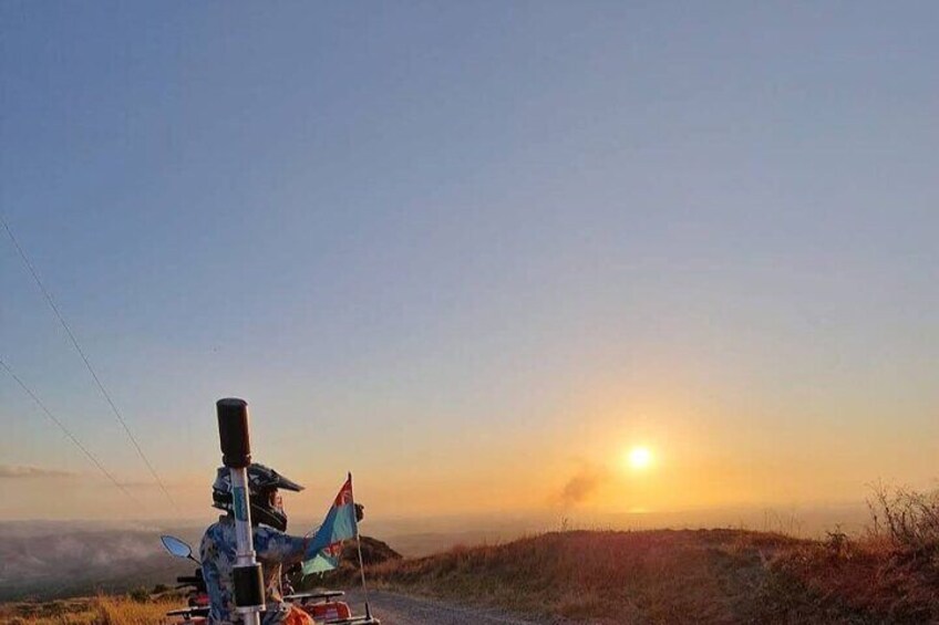 Een zonsondergang boven een glooiend landschap, met een motorfiets en een vlag aan de linkerkant. 1