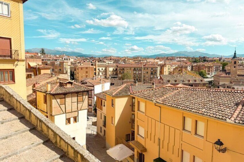 A cityscape with terracotta-roofed buildings, a clear sky, and distant mountains. 3