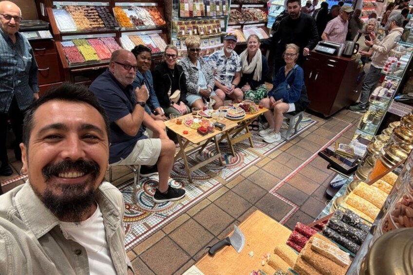 Un gruppo di persone sedute a un tavolo in un mercato, con diversi prodotti alimentari esposti sullo sfondo. 4