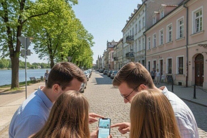 A group of people looking at a smartphone on a cobblestone street with buildings and a river in the background. 2