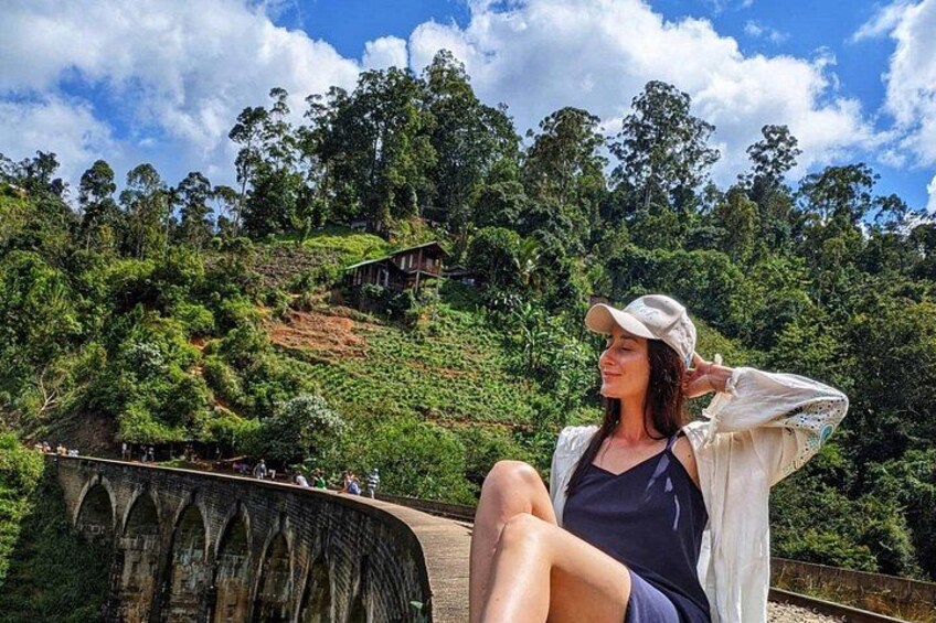 Una persona sentada en un puente, con un paisaje verde exuberante y un puente de piedra al fondo. 2