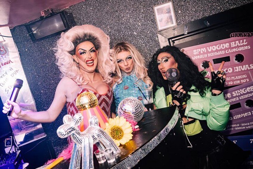 Drei Personen in einem Raum mit glitzernden Wänden, einem Mikrofon und einem Schild mit der Aufschrift '8PM VOGUE BALL'. 1