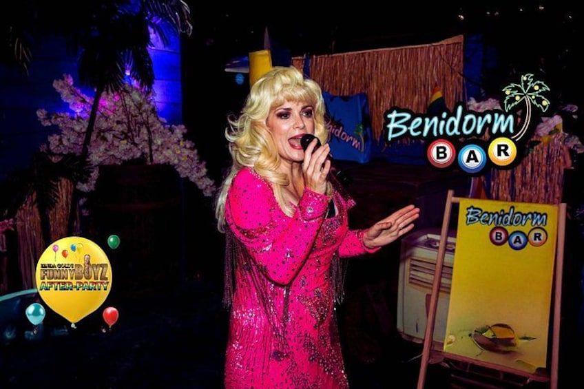 Eine Person in einem pinkfarbenen, pailletenbesetzten Outfit im Benidorm Bar, mit tropischem Hintergrund inklusive Palmen und einer Strandansicht. 5