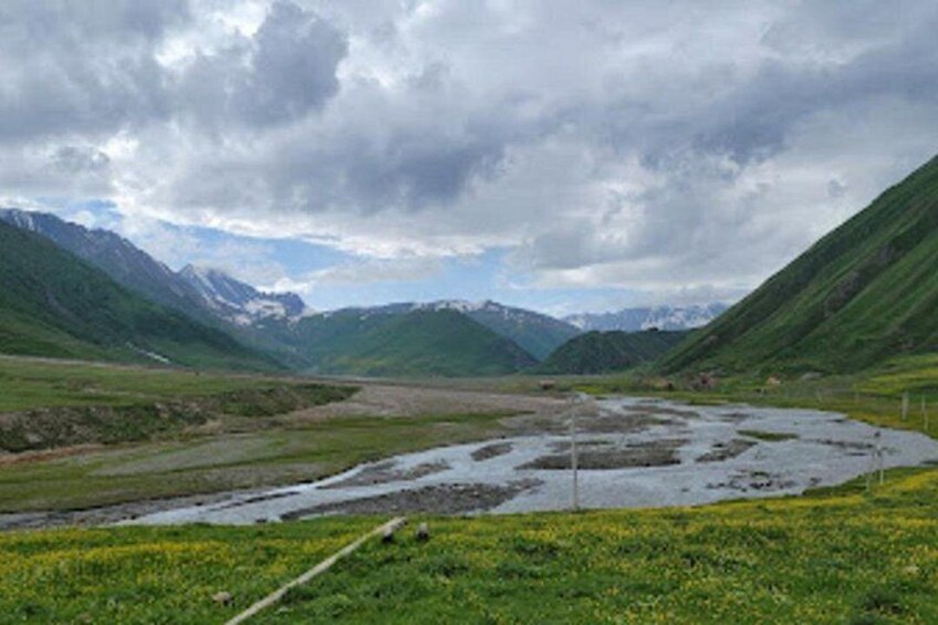 Eine gebirgige Landschaft mit einem Fluss, grünen Feldern und fernen, schneebedeckten Gipfeln unter einem bewölkten Himmel. 2