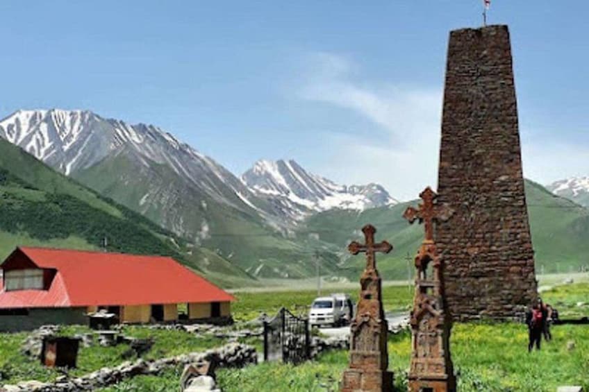 Ein Steinkreuz, ein Gebäude mit rotem Dach und verschneite Berge im Hintergrund. 1