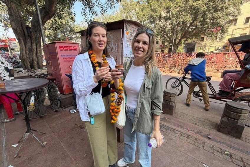 Two people standing outdoors, one holding a drink and the other holding a camera. There is a bicycle and a rickshaw in the background. 5