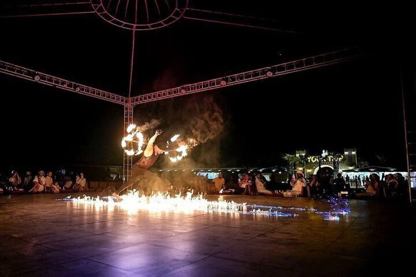 A nighttime outdoor performance with fire and smoke effects, a crowd of spectators, and a stage with a metal framework. 3