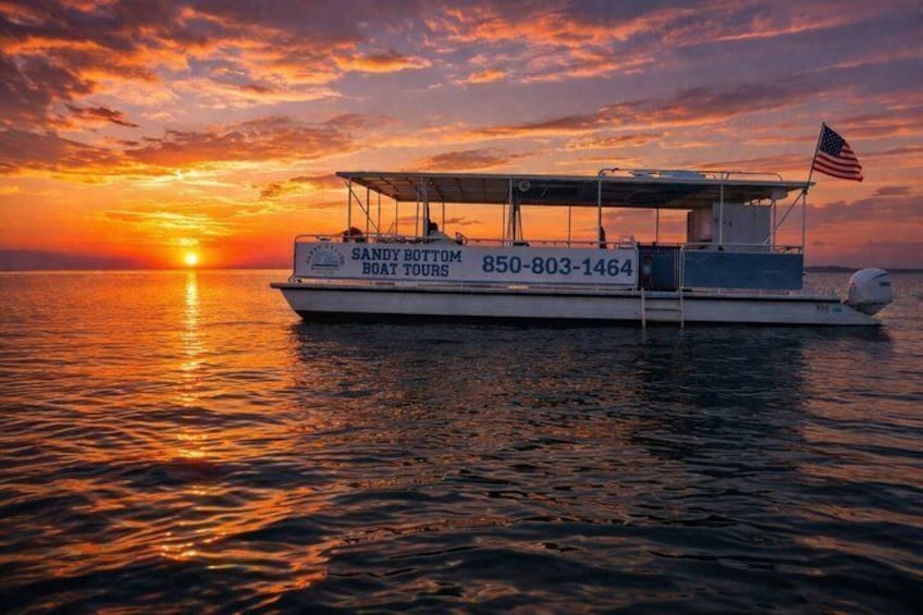 Un bateau portant l’inscription « Sandy Bottom Boat Tours » et arborant un numéro de téléphone sur sa coque, naviguant sur une étendue d’eau au coucher du soleil. 1