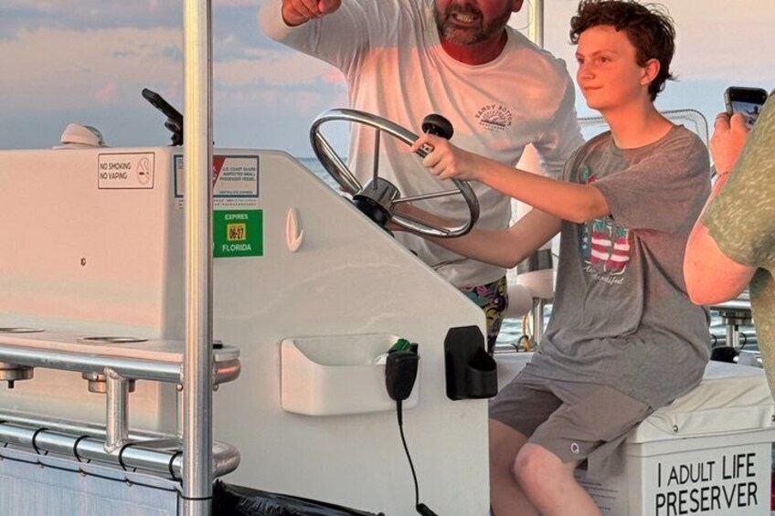 Une personne tenant un volant sur un bateau, une autre personne tenant un microphone, et une pancarte sur laquelle on peut lire : « Gilet de sauvetage pour adultes ». 4