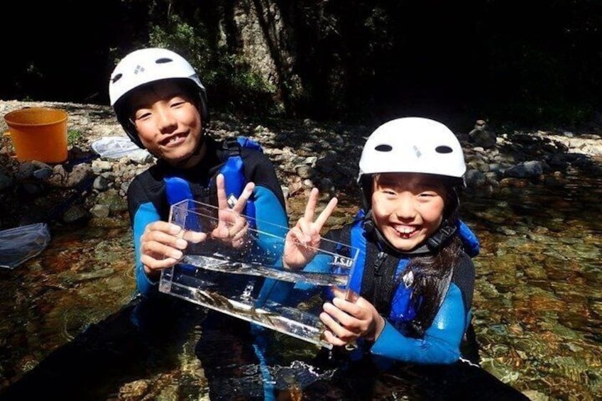 Two people wearing helmets and life jackets, holding a transparent object, in a rocky outdoor setting. 3