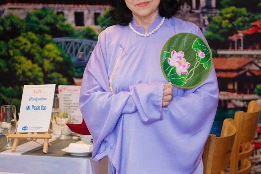 Eine Person hält einen grün bestickten Fächer mit rosa Blumen und steht an einem Tisch, auf dem ein Schild mit dem Text ‚Ms Thanh Van‘ steht. 2