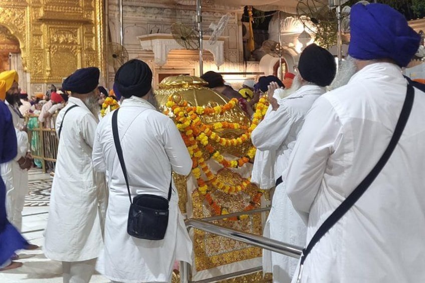 A group of people in white robes and turbans, standing in a temple with golden decorations and floral arrangements. 1
