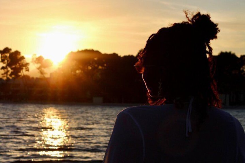A silhouette of a person with a bun hairstyle, wearing a white top, standing by a body of water during sunset. 5
