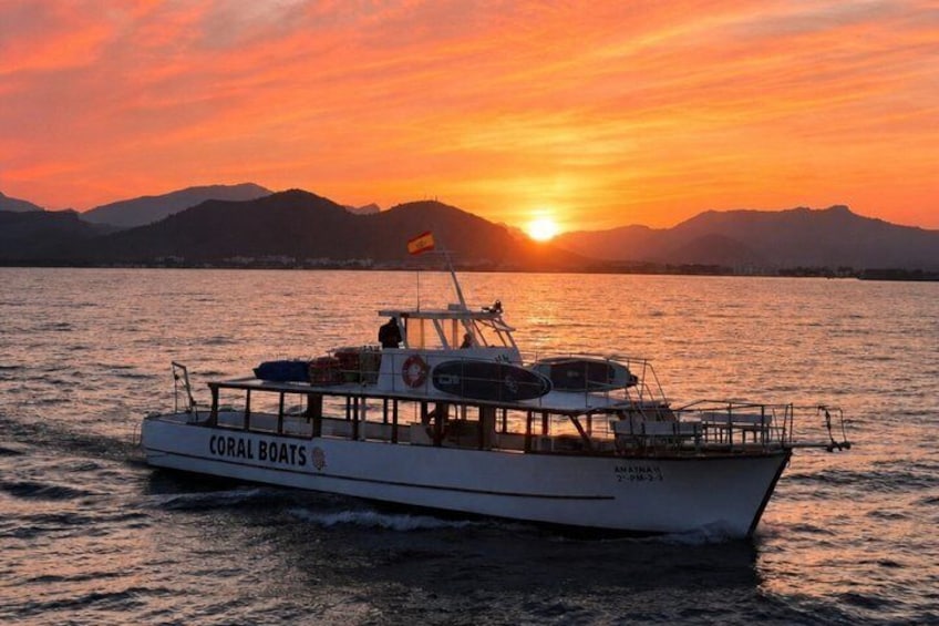 A boat named 'CORAL BOATS' with a flag, on calm water during sunset with mountains in the background. 1