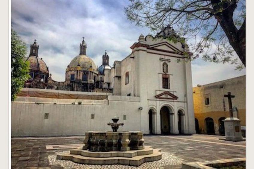 A courtyard with a fountain, a church with domes, and a cross. 2