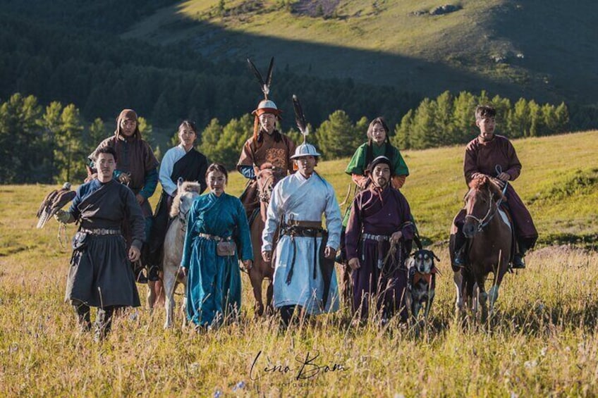 A group of people in traditional attire standing in a grassy field with a mountainous background. 1