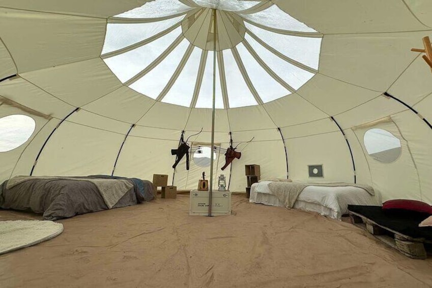 A large, round, white tent with a central skylight. Inside, there are two beds with white bedding and a small table. 2