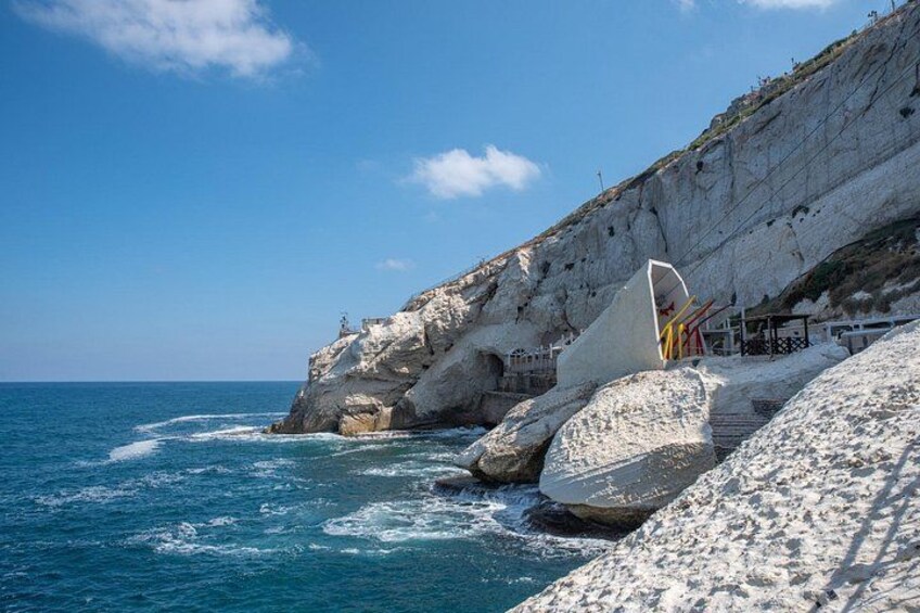 A rocky coastline with waves crashing against the shore, a white cliff, and a structure with a curved roof. 1