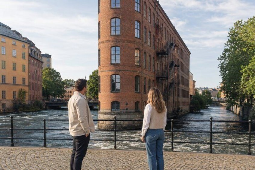 Two people standing on a cobblestone path by a river, with a brick building and other buildings in the background. 4
