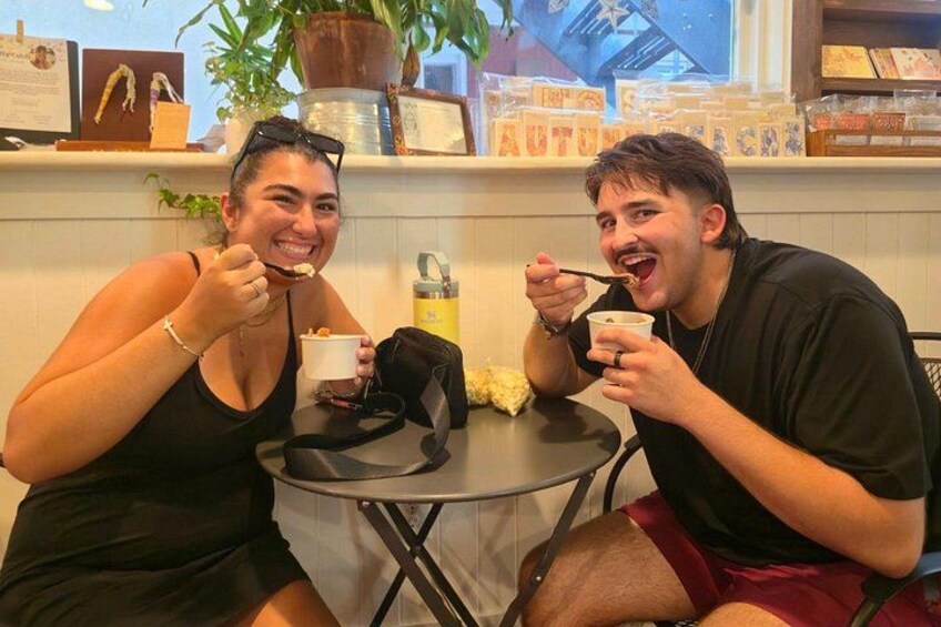 Two people sitting at a table with cups, a bag, and a plant in the background. 1