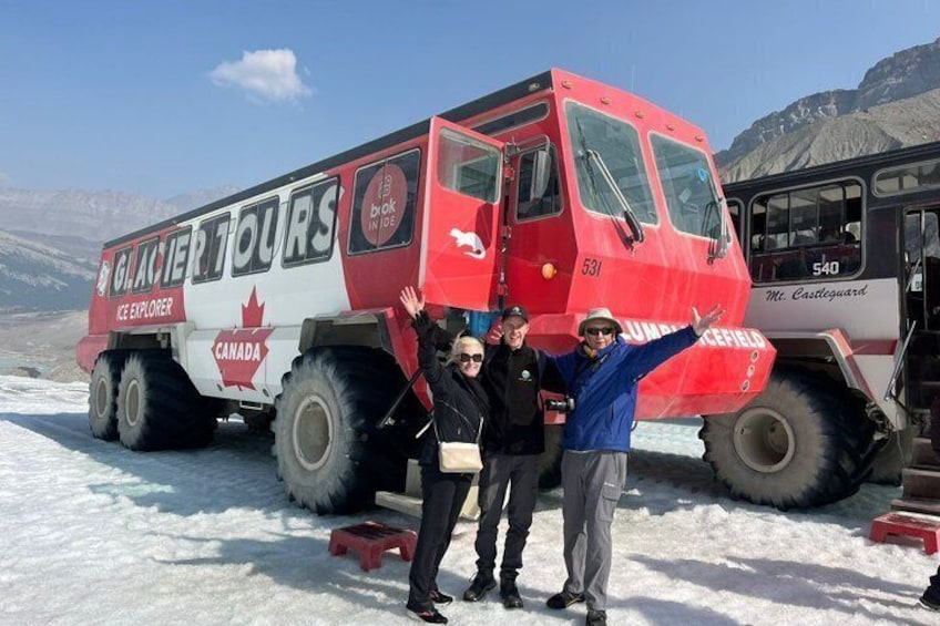 Een rode en witte toeristenbus met 'Glacier Tours' en 'Canada' erop, geparkeerd in de sneeuw met bergen op de achtergrond. 1