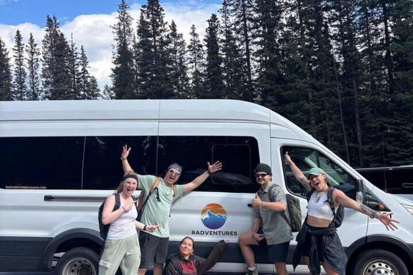 Een groep mensen poseert voor een witte bus met een logo, omgeven door een bos. 2
