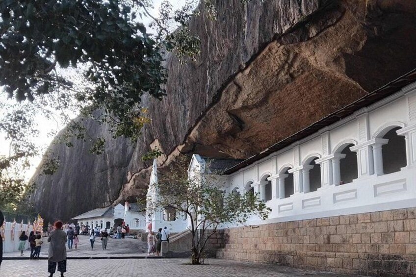 A rock formation with carved figures, a white building with arched windows, and a paved area with people. 3