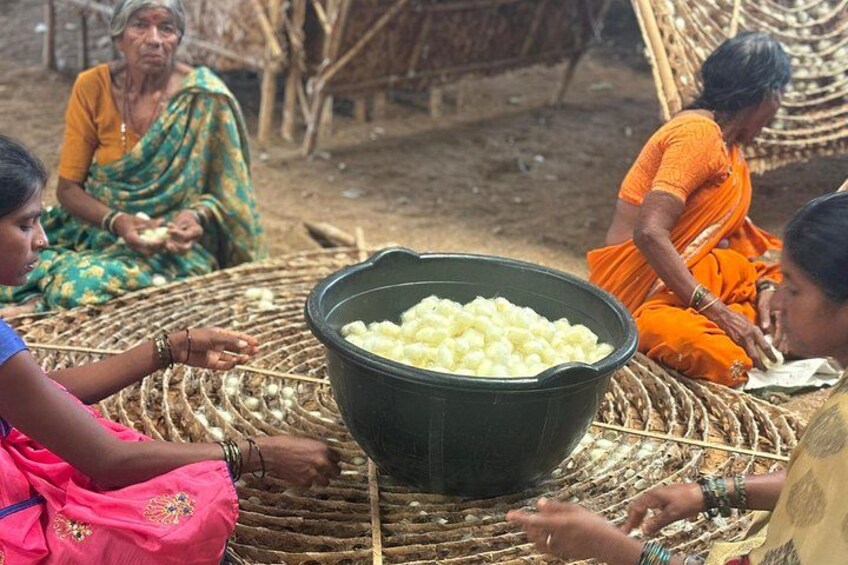 Women sitting around a woven mat, with a large black bowl of white items in the center. 4
