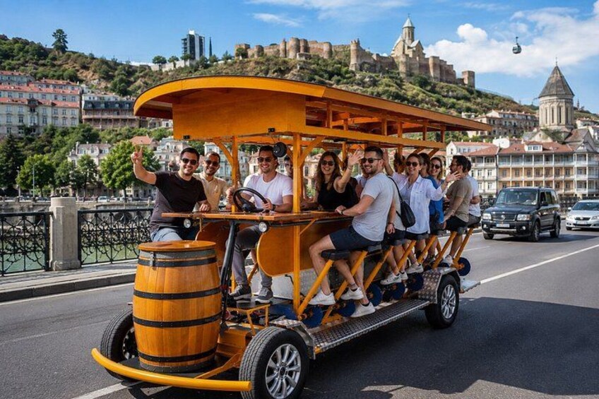 A group of people on a wooden trolley with a barrel on the back, driving on a road with a cityscape and a hill with a castle in the background. 2