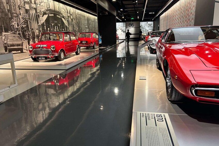 A museum exhibit with vintage cars, including a red sports car and a red van, displayed on a reflective floor with a cityscape mural in the background. 1