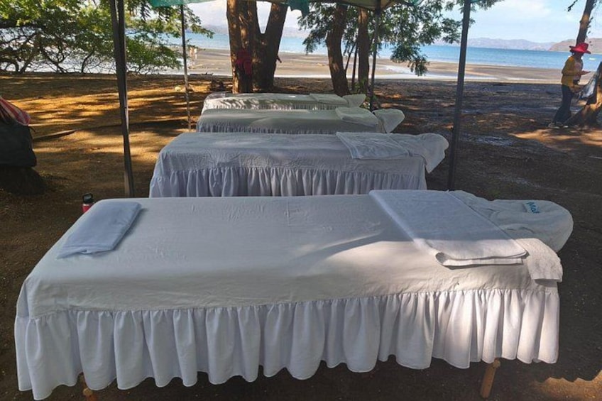 A row of white beds with white sheets and pillows, set up outdoors with trees and a beach in the background. 1
