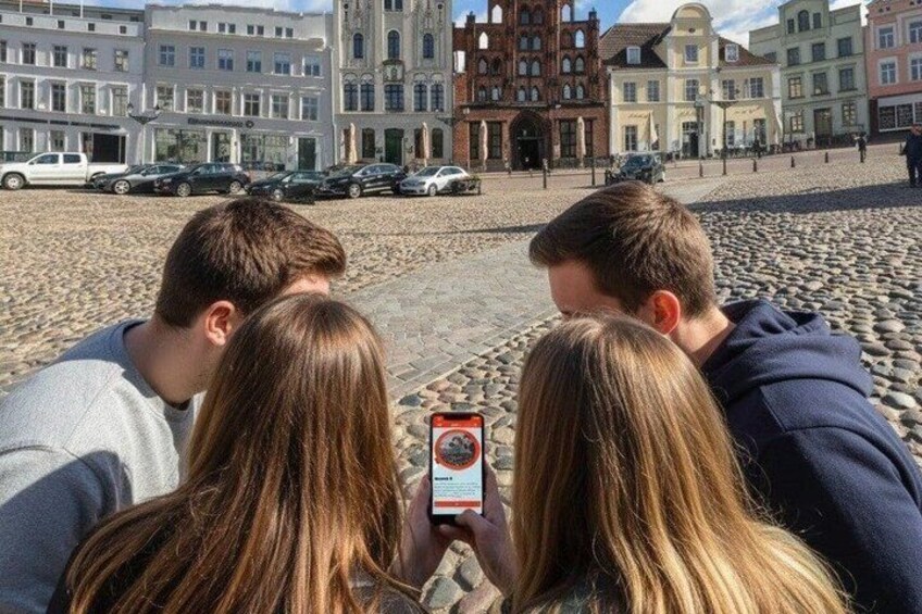 Three people taking a photo with a smartphone in a cobblestone square with historic buildings in the background. 4