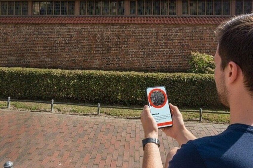 A person holding a smartphone with a red circle and a black center, standing on a brick-paved area with a brick wall and a hedge in the background. 3