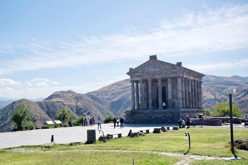A classical building with columns, surrounded by a grassy area and mountains in the background. 1